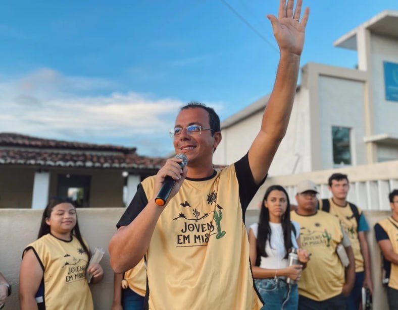 Missionário de colete amarelo com o braço levantado e o microfone, com a equipe ao fundo